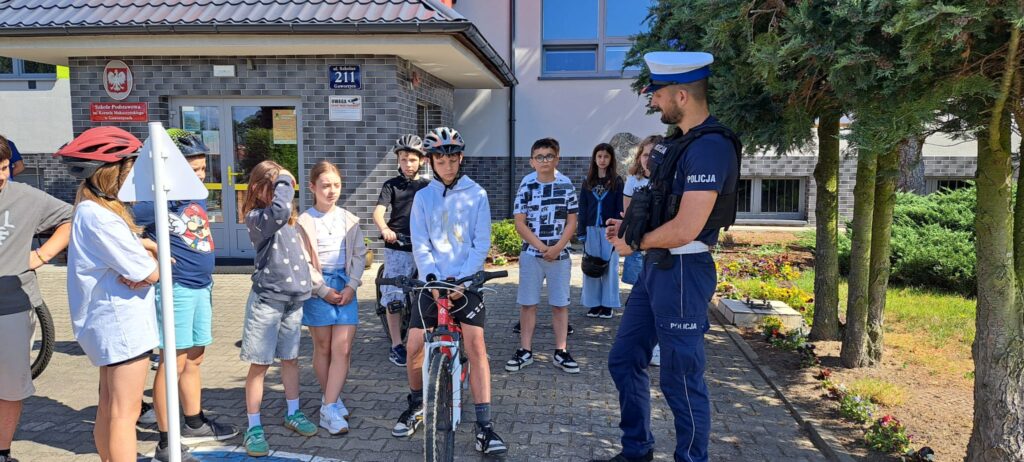 Na zdjęciu widać grupę dzieci stojących przed budynkiem szkoły podczas zajęć związanych z bezpieczeństwem ruchu drogowego. Przed nimi stoi umundurowany policjant, który prawdopodobnie prowadzi pogadankę lub instruktaż.
Kilkoro uczniów ma na głowach kaski rowerowe, a jeden z chłopców siedzi na rowerze, co sugeruje, że spotkanie dotyczy bezpiecznej jazdy na rowerze. Dzieci słuchają funkcjonariusza z uwagą, stojąc na szkolnym dziedzińcu. W tle znajduje się wejście do szkoły z tablicami informacyjnymi oraz zadbany teren z kwiatami i drzewami.