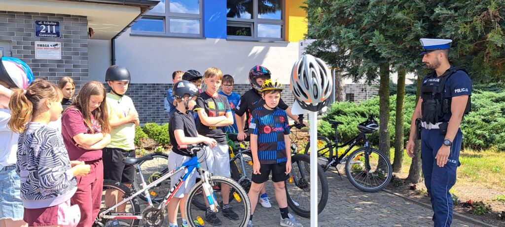 Na zdjęciu przedstawiono grupę uczniów uczestniczących w zajęciach dotyczących bezpieczeństwa ruchu drogowego. Dzieci stoją przed budynkiem szkoły, obok swoich rowerów, i z uwagą słuchają policjanta prowadzącego spotkanie edukacyjne.
Większość uczniów ma na głowach kaski rowerowe, a w centralnej części fotografii znajduje się duży model kasku umieszczony na stojaku, który stanowi pomoc dydaktyczną podczas omawiania zasad bezpiecznej jazdy. Policjant w umundurowaniu wyjaśnia uczestnikom znaczenie stosowania kasków ochronnych oraz przestrzegania przepisów ruchu drogowego.