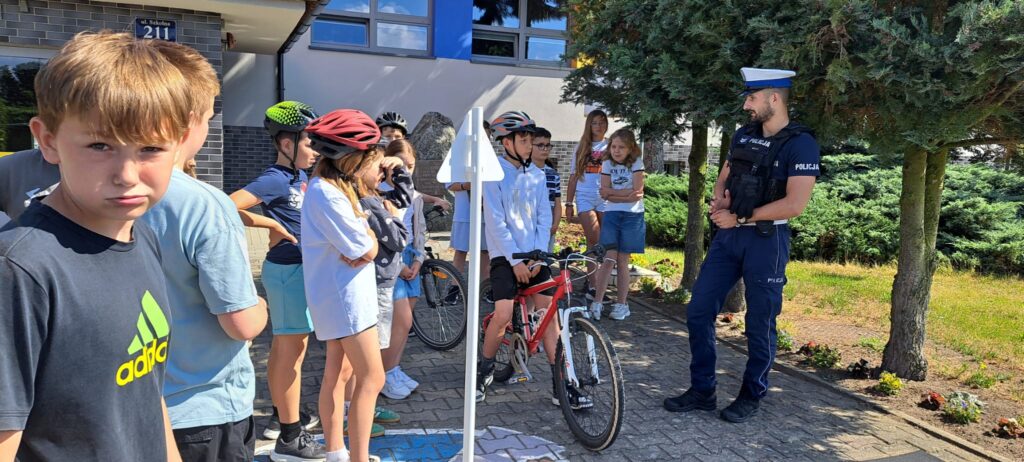 a zdjęciu przedstawiono grupę uczniów uczestniczących w spotkaniu edukacyjnym dotyczącym bezpieczeństwa ruchu drogowego. Dzieci zgromadziły się przed budynkiem szkoły, gdzie funkcjonariusz policji prowadzi pogadankę na temat zasad bezpiecznego poruszania się po drogach.
W centrum grupy znajduje się chłopiec siedzący na czerwonym rowerze, a wokół niego stoją pozostali uczniowie, z których część ma założone kaski rowerowe. Na pierwszym planie, po lewej stronie, widoczny jest chłopiec patrzący w stronę obiektywu. Policjant, stojący po prawej stronie zdjęcia, zwraca się do dzieci, wyjaśniając zagadnienia związane z bezpieczeństwem rowerzystów.