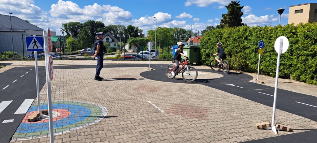 Na zdjęciu przedstawiono szkolne miasteczko ruchu drogowego, na którym odbywają się praktyczne zajęcia dla dzieci z zakresu bezpiecznego poruszania się rowerem. W centralnej części fotografii stoi policjant, który nadzoruje przebieg ćwiczeń i obserwuje uczestników.
Dwoje uczniów w kaskach rowerowych porusza się po wyznaczonej trasie, ćwicząc jazdę zgodnie z zasadami ruchu drogowego. Jeden z rowerzystów jedzie po rondzie, a drugi zbliża się do niego, stosując się do obowiązującego oznakowania.