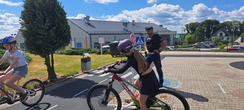 Na zdjęciu przedstawiono zajęcia praktyczne z zakresu bezpieczeństwa ruchu drogowego prowadzone na szkolnym miasteczku rowerowym. Dwie uczennice w kaskach ochronnych poruszają się na rowerach po wyznaczonej trasie, ćwicząc prawidłowe zachowania na drodze.
W centrum fotografii znajduje się dziewczynka przygotowująca się do włączenia do ruchu na oznakowanej jezdni, natomiast po lewej stronie druga uczestniczka jedzie rowerem zgodnie z wyznaczonym kierunkiem. Nad przebiegiem ćwiczeń czuwa policjant, który obserwuje młodych rowerzystów i ocenia poprawność wykonywanych manewrów.