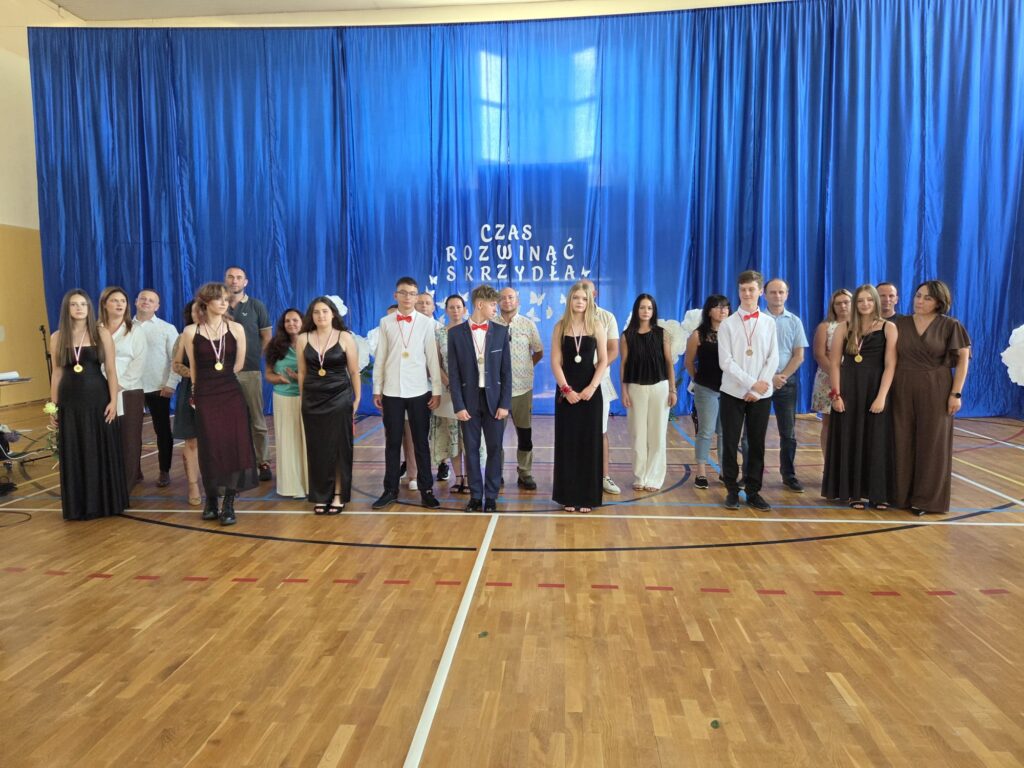 Na zdjęciu przedstawiono pamiątkową fotografię absolwentów wraz z rodzicami oraz wychowawczynią, wykonaną podczas uroczystości zakończenia roku szkolnego. Wszyscy uczestnicy ustawili się na środku szkolnej sali gimnastycznej na tle niebieskiej kurtyny z napisem „Czas rozwinąć skrzydła”, symbolizującym zakończenie ważnego etapu edukacji i rozpoczęcie nowych życiowych wyzwań.

W pierwszym rzędzie stoją absolwenci ubrani w eleganckie stroje galowe. Dziewczęta mają na sobie długie czarne lub ciemne suknie, natomiast chłopcy występują w białych koszulach z czerwonymi muszkami lub w garniturach. Na szyjach wielu uczniów widoczne są medale, będące wyróżnieniem za osiągnięcia i zaangażowanie podczas nauki.

Za absolwentami stoją rodzice i opiekunowie, którzy wspólnie z dziećmi pozują do pamiątkowego zdjęcia. Ich obecność podkreśla wagę wydarzenia oraz wsparcie, jakie towarzyszyło uczniom przez cały okres edukacji. Po prawej stronie fotografii znajduje się wychowawczyni klasy, ubrana w elegancką brązową suknię, stojąca obok swoich uczniów.