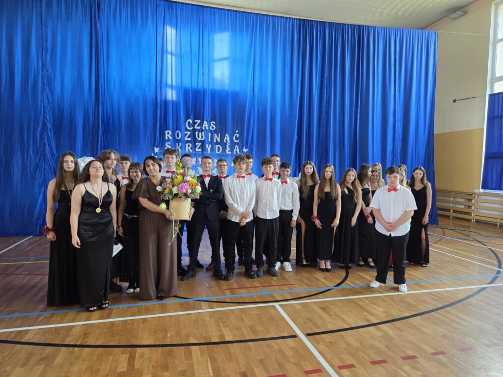 Na zdjęciu przedstawiono grupową fotografię wykonaną podczas uroczystości zakończenia roku szkolnego i pożegnania absolwentów. W centrum kadru stoją uczniowie klasy kończącej szkołę wraz z wychowawczynią, pozując do wspólnego pamiątkowego zdjęcia.

Uczniowie ubrani są w eleganckie stroje galowe. Dziewczęta mają na sobie długie czarne suknie, a chłopcy występują w białych koszulach z czerwonymi muszkami lub ciemnych garniturach. W pierwszym rzędzie, po lewej stronie, jedna z uczennic ma zawieszony na szyi medal. Na środku grupy stoi wychowawczyni w długiej brązowej sukni, trzymająca okazały bukiet kolorowych kwiatów – wyraz wdzięczności i podziękowania od uczniów za wspólnie spędzone lata.