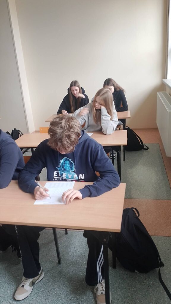 Na zdjęciu widać salę lekcyjną podczas pisania sprawdzianu lub testu. Na pierwszym planie siedzi uczeń w granatowej bluzie z kapturem, pochylony nad kartką i wypełniający arkusz. W tle przy kolejnych ławkach siedzą trzy uczennice – dwie z nich również pracują nad zadaniami, a jedna opiera głowę na ręce, skupiając się na swojej pracy.