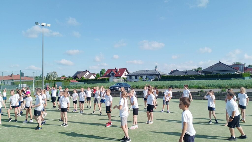Na zdjęciu widać dużą grupę dzieci uczestniczących w zajęciach sportowych na szkolnym boisku wielofunkcyjnym. Uczniowie są ubrani w białe koszulki i sportowe spodenki, a większość z nich wykonuje jednocześnie te same ruchy, co sugeruje rozgrzewkę, ćwiczenia koordynacyjne lub wspólny trening.

Boisko znajduje się na świeżym powietrzu w słoneczny dzień. Nad uczestnikami rozciąga się błękitne niebo z niewielką liczbą chmur. Długie cienie rzucane przez dzieci wskazują, że zdjęcie zostało wykonane rano lub późnym popołudniem.