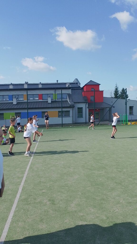 Na zdjęciu widać uczniów uczestniczących w zajęciach sportowych na szkolnym boisku. Kilkoro dzieci stoi w różnych miejscach na wyznaczonym polu gry, przyjmując pozycje charakterystyczne dla gry zespołowej, prawdopodobnie siatkówki, „zbijaka” lub innej zabawy ruchowej wymagającej współpracy i szybkiej reakcji.

Uczniowie ubrani są w stroje sportowe – białe koszulki, krótkie spodenki i obuwie sportowe. Niektórzy obserwują przebieg gry, inni są gotowi do przyjęcia piłki lub wykonania kolejnego ruchu. W tle widać również grupę dzieci siedzących przy ogrodzeniu boiska, które prawdopodobnie czekają na swoją kolej lub kibicują uczestnikom.