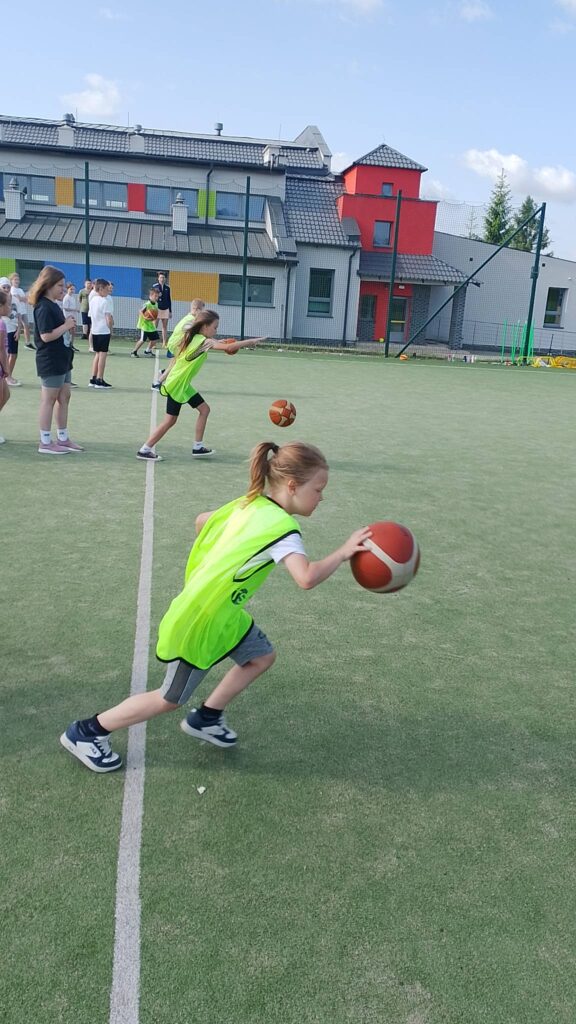 Na zdjęciu przedstawiono zajęcia sportowe odbywające się na szkolnym boisku. Na pierwszym planie widać dziewczynkę w jaskrawozielonej kamizelce sportowej, która wykonuje kozłowanie piłką do koszykówki. Jest pochylona do przodu i skupiona na ćwiczeniu, co sugeruje udział w treningu lub konkurencji sprawnościowej.

W tle znajdują się inni uczniowie, również ubrani w stroje sportowe. Część z nich obserwuje przebieg ćwiczenia, a niektórzy biorą udział w podobnych zadaniach z piłkami. Atmosfera wskazuje na zajęcia wychowania fizycznego, szkolny turniej lub dzień sportu.