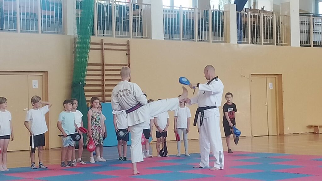 Na zdjęciu widać zajęcia sportowe lub pokaz sztuk walki odbywający się w sali gimnastycznej. Na pierwszym planie dwóch instruktorów ubranych w białe stroje do taekwondo demonstruje technikę kopnięcia. Jeden z nich wykonuje wysokie kopnięcie, trafiając stopą w specjalną tarczę treningową trzymaną przez drugiego instruktora.

Wokół nich stoją dzieci, które obserwują pokaz. Część uczestników trzyma w rękach kolorowe tarcze do ćwiczeń, co sugeruje, że biorą udział w treningu lub warsztatach. Dzieci są ubrane w stroje sportowe, głównie jasne koszulki i krótkie spodenki.