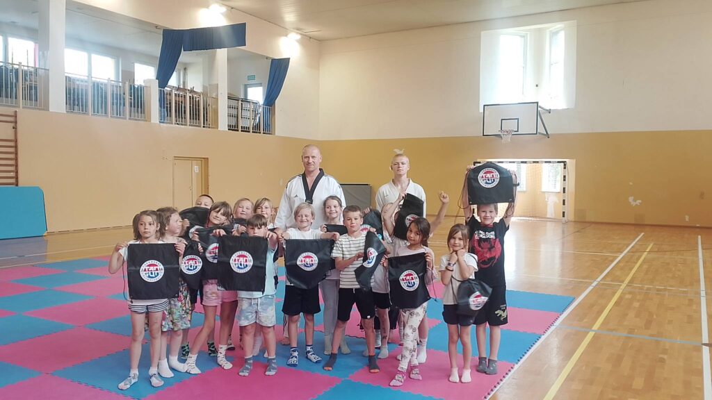 Na zdjęciu znajduje się grupa dzieci oraz dwóch instruktorów pozujących do pamiątkowej fotografii w sali gimnastycznej po zakończonych zajęciach sportowych lub warsztatach taekwondo. Dzieci stoją na kolorowych matach treningowych w kolorze różowym i niebieskim, trzymając w rękach czarne worki lub plecaki z logo klubu, które prawdopodobnie otrzymały jako upominki lub nagrody za udział w zajęciach.
W centrum grupy stoją dwaj instruktorzy ubrani w tradycyjne stroje do taekwondo