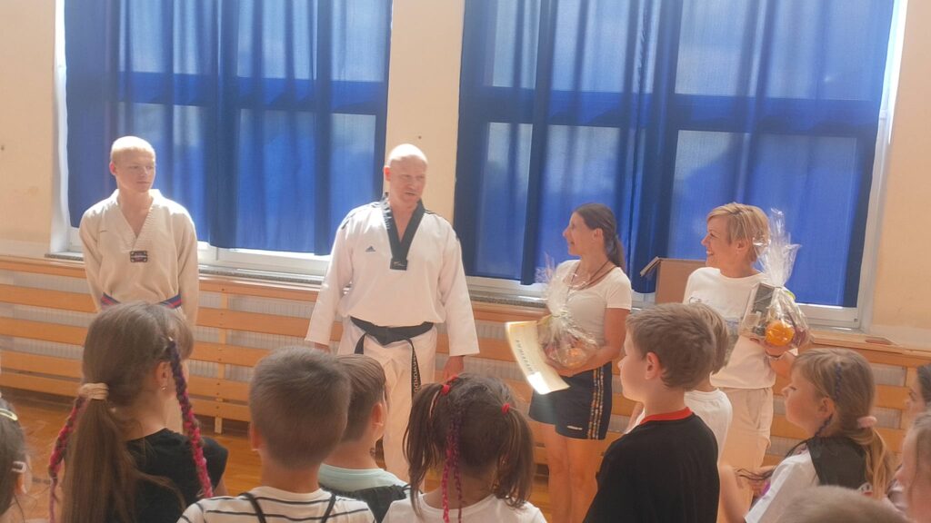 Na zdjęciu widać zakończenie zajęć sportowych lub spotkania związanego z taekwondo w sali gimnastycznej. Na pierwszym planie stoją dzieci, które z zainteresowaniem słuchają osób znajdujących się przed nimi.

W centrum fotografii znajduje się dwóch instruktorów ubranych w tradycyjne stroje do taekwondo. Obok nich stoją dwie kobiety trzymające ozdobnie zapakowane upominki lub kosze prezentowe. Jedna z kobiet trzyma również dokument lub dyplom. Wszyscy wyglądają na zadowolonych i zaangażowanych w uroczyste podsumowanie wydarzenia.