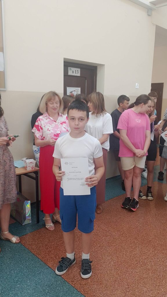 Na zdjęciu widać ucznia stojącego na szkolnym korytarzu podczas uroczystego apelu podsumowującego konkursy i osiągnięcia uczniów. Chłopiec trzyma w rękach dyplom, który otrzymał za udział lub sukces w konkursie. Ubrany jest w białą koszulkę, niebieskie spodenki i sportowe buty.

W tle znajdują się nauczyciele oraz uczniowie oczekujący na wręczenie nagród lub zakończenie uroczystości. Obok stoi stolik z przygotowanymi upominkami i materiałami związanymi z wydarzeniem. Atmosfera jest oficjalna, ale jednocześnie radosna i pełna dumy z osiągnięć uczniów