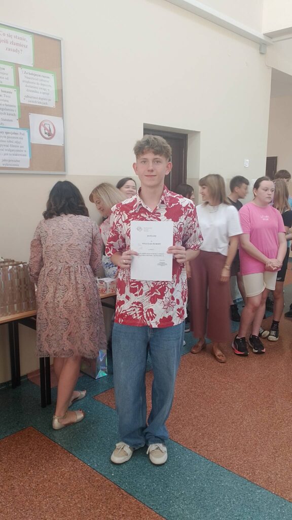 Na zdjęciu widoczny jest uczeń stojący w szkolnym korytarzu podczas uroczystego apelu. Trzyma w rękach dyplom otrzymany za osiągnięcia w konkursie szkolnym. Ubrany jest w czerwoną koszulę we wzory oraz jasne jeansy. W tle znajdują się nauczyciele i uczniowie uczestniczący w ceremonii wręczania nagród. Po lewej stronie widoczny jest stolik z upominkami i nagrodami przygotowanymi dla laureatów konkursów. Atmosfera wydarzenia jest uroczysta i radosna, a zdjęcie dokumentuje moment wyróżnienia ucznia za jego sukcesy w II semestrze roku szkolnego.
