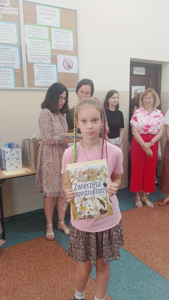 Na zdjęciu widoczna jest uczennica stojąca na szkolnym korytarzu podczas uroczystego apelu podsumowującego konkursy szkolne. Dziewczynka trzyma w rękach książkę zatytułowaną „Zwierzęta konstruktorzy”, która została wręczona jej jako nagroda za osiągnięcia w konkursie. Ubrana jest w różową koszulkę i wzorzystą spódnicę, a jej kolorowe warkoczyki dodają fotografii radosnego charakteru.

W tle znajdują się nauczyciele przygotowujący nagrody oraz inni uczestnicy uroczystości. Na stoliku widoczne są upominki i pakunki przeznaczone dla laureatów konkursów. Zdjęcie przedstawia moment wyróżnienia uczennicy i stanowi pamiątkę jej sukcesu oraz aktywnego udziału w życiu szkoły. 