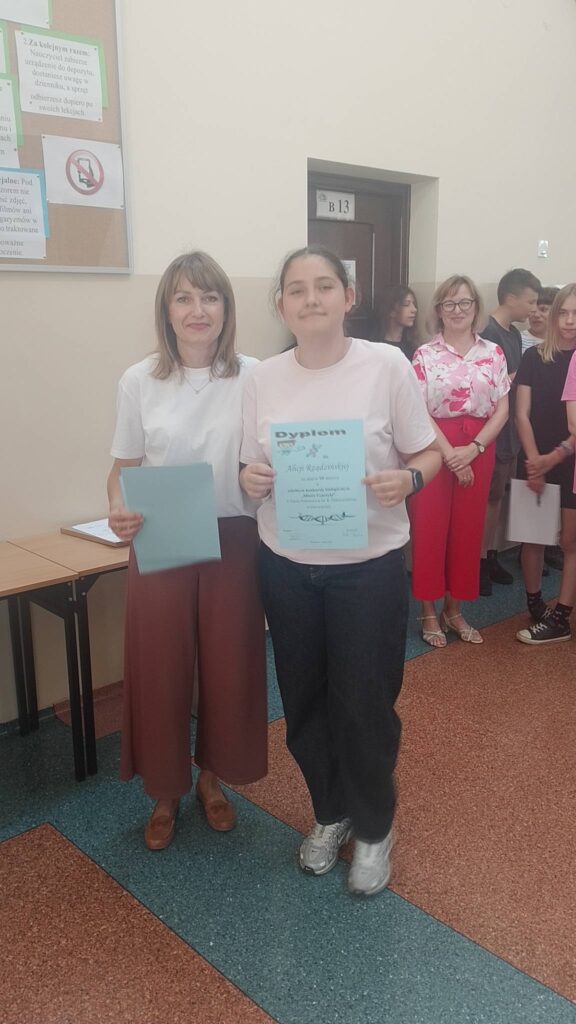 Na zdjęciu uwieczniono moment wręczenia nagrody podczas szkolnego apelu podsumowującego konkursy i osiągnięcia uczniów w II semestrze. Na pierwszym planie znajduje się uczennica trzymająca dyplom, stojąca obok nauczycielki. Obydwie pozują do pamiątkowego zdjęcia i uśmiechają się do aparatu.

W tle widoczni są inni uczniowie oraz nauczyciele uczestniczący w uroczystości. Niektórzy trzymają dyplomy i oczekują na swoje wyróżnienia. Zdjęcie zostało wykonane na szkolnym korytarzu, gdzie odbywało się wręczanie nagród. Panuje uroczysta i serdeczna atmosfera, podkreślająca sukcesy uczniów oraz ich zaangażowanie w konkursy i życie szkoły.