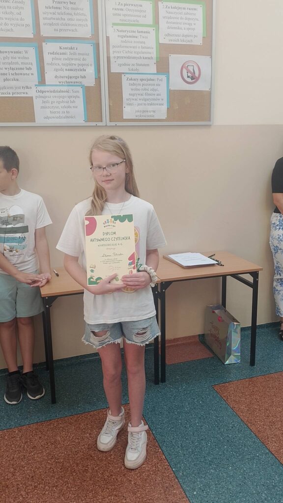 Na zdjęciu widać uczennicę stojącą w sali szkolnej podczas uroczystości lub podsumowania konkursu. Dziewczynka trzyma w rękach dyplom z napisem „Dyplom Aktywnego Czytelnika”, co sugeruje, że została wyróżniona za zaangażowanie w czytanie książek.

Ma na sobie białą koszulkę, jasne dżinsowe szorty oraz białe sportowe buty. Nosi okulary i ma długie, jasne włosy. Patrzy w stronę aparatu z lekkim, spokojnym uśmiechem.

W tle znajdują się szkolne ławki oraz tablica korkowa z regulaminami i informacjami dotyczącymi korzystania z telefonów komórkowych w szkole.