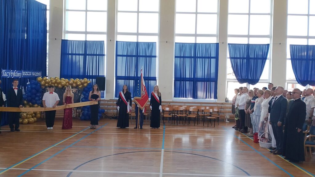 Na zdjęciu przedstawiono uroczystą akademię z okazji zakończenia roku szkolnego i pożegnania absolwentów, odbywającą się w szkolnej sali gimnastycznej. Fotografia ukazuje szeroki plan sali, dzięki czemu widoczni są zarówno uczniowie, jak i zaproszeni goście.W centralnej części zdjęcia znajduje się poczet sztandarowy. Uczniowie ubrani w galowe stroje z biało-czerwonymi szarfami stoją na baczność przy sztandarze szkoły, nadając uroczystości podniosły charakter

Po lewej stronie stoją uczniowie ubrani w stroje galowe. Chłopcy mają na sobie białe koszule z czerwonymi muszkami lub ciemne garnitury, natomiast dziewczęta występują w eleganckich, długich sukniach.