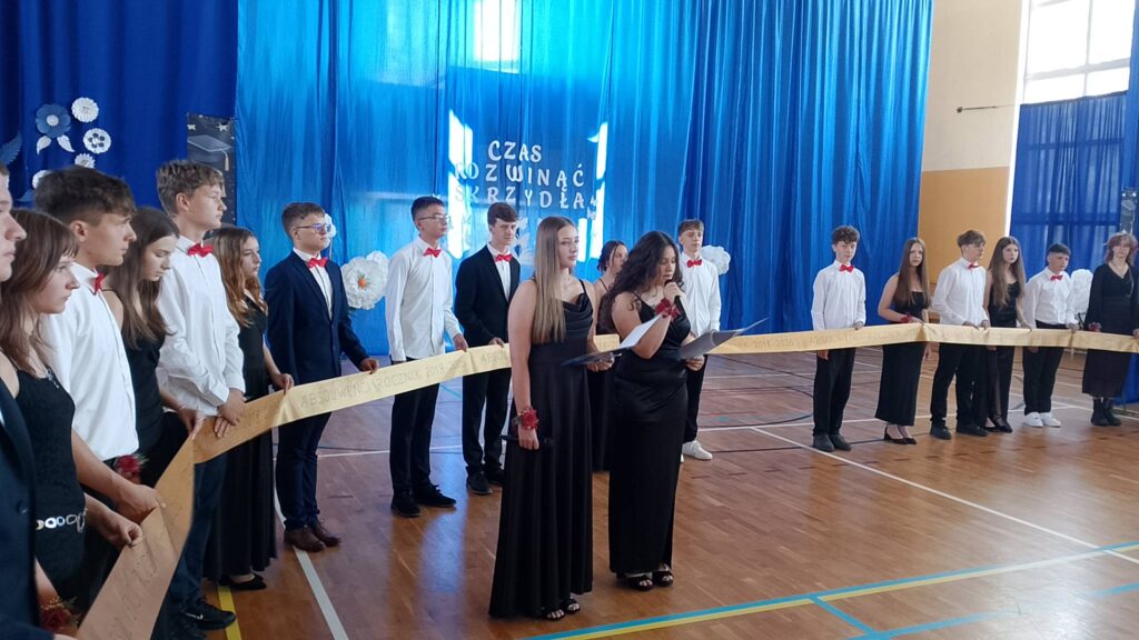 Na zdjęciu przedstawiono szeroki plan uroczystości zakończenia roku szkolnego i pożegnania absolwentów, odbywającej się w sali gimnastycznej udekorowanej na tę okazję. W centrum wydarzenia znajduje się grupa uczniów ubranych w eleganckie stroje galowe – chłopcy mają na sobie białe koszule, czarne spodnie i czerwone muszki lub garnitury, natomiast dziewczęta występują w długich, wizytowych sukniach.

Uczniowie stoją w półkolu, wspólnie trzymając długą, złotą wstęgę z napisem „Absolwenci Rocznik 2018–2026”, która symbolicznie podkreśla zakończenie nauki w szkole podstawowej. W centralnej części kadru dwie uczennice stoją przy mikrofonach i czytają tekst z przygotowanych kartek, prowadząc oficjalną część akademii.
