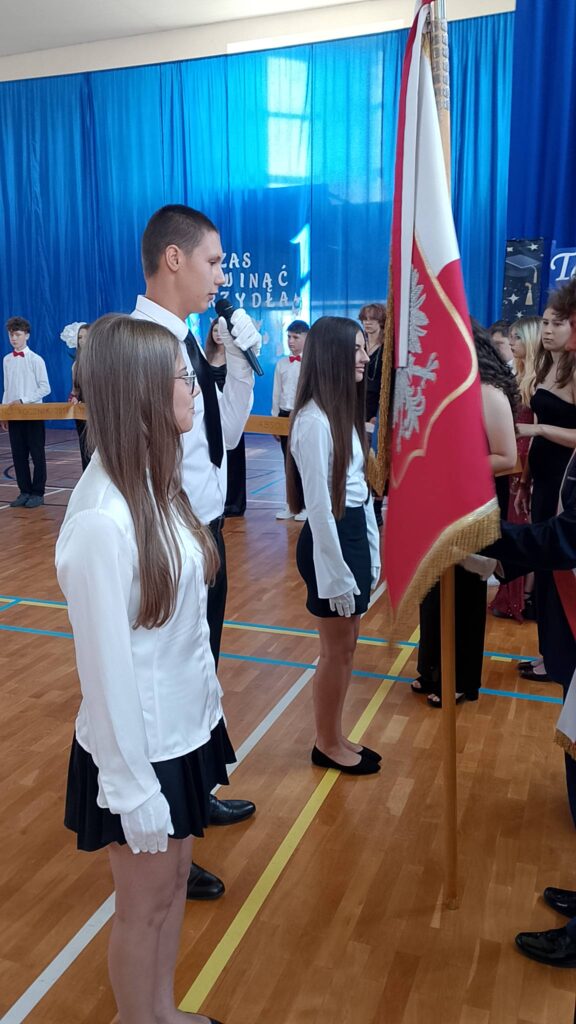 Na zdjęciu przedstawiono uroczysty moment przekazania sztandaru podczas szkolnej akademii związanej z zakończeniem roku szkolnego i pożegnaniem absolwentów. W centrum kadru znajduje się poczet sztandarowy. Uczniowie ubrani są w galowe stroje – białe koszule, czarne spódnice lub spodnie, białe rękawiczki oraz biało-czerwone szarfy. Jedna z uczennic trzyma sztandar szkoły z czerwonym płatem ozdobionym białym orłem.

Obok sztandaru stoi uczeń z mikrofonem, który odczytuje tekst związany z ceremonią przekazania sztandaru. Pozostali członkowie pocztu zachowują postawę na baczność, podkreślając uroczysty charakter wydarzenia.
