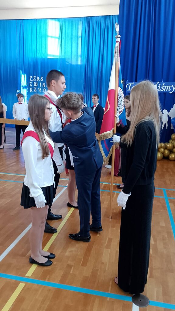 Na zdjęciu przedstawiono uroczysty moment przekazania pocztu sztandarowego podczas szkolnej akademii z okazji zakończenia roku szkolnego. W centrum kadru znajdują się członkowie ustępującego i nowego pocztu sztandarowego, ubrani w stroje galowe. Uczniowie mają na sobie białe koszule, czarne spódnice lub spodnie, białe rękawiczki oraz biało-czerwone szarfy, które podkreślają ceremonialny charakter wydarzenia.

W centralnej części fotografii widoczny jest uczeń w granatowym garniturze, który pomaga założyć biało-czerwoną szarfę członkowi nowego pocztu sztandarowego. Obok stoi uczennica trzymająca sztandar szkoły z biało-czerwonym płatem i godłem państwowym. Pozostali uczestnicy ceremonii zachowują postawę wyprostowaną i z powagą obserwują przebieg uroczystości.