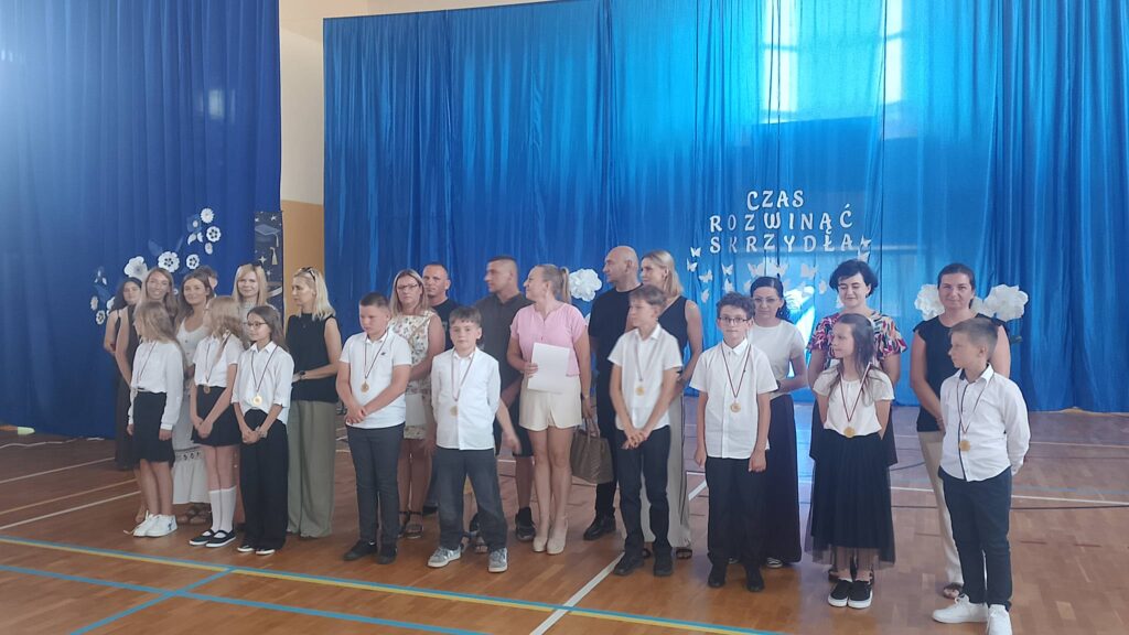 Na zdjęciu przedstawiono grupę uczniów wraz z rodzicami podczas uroczystości zakończenia roku szkolnego w szkolnej sali gimnastycznej. W pierwszym rzędzie stoją uczniowie ubrani w odświętne stroje – białe koszule i bluzki oraz ciemne spodnie lub spódnice. Na szyjach mają zawieszone medale, które świadczą o otrzymanych wyróżnieniach i osiągnięciach.

Za dziećmi stoją rodzice i opiekunowie, którzy uczestniczą w uroczystości. Ubrani są elegancko, a ich obecność podkreśla wyjątkowy charakter wydarzenia oraz wspólne przeżywanie sukcesów uczniów. Wszyscy zwróceni są w stronę fotografa i pozują do pamiątkowego zdjęcia.