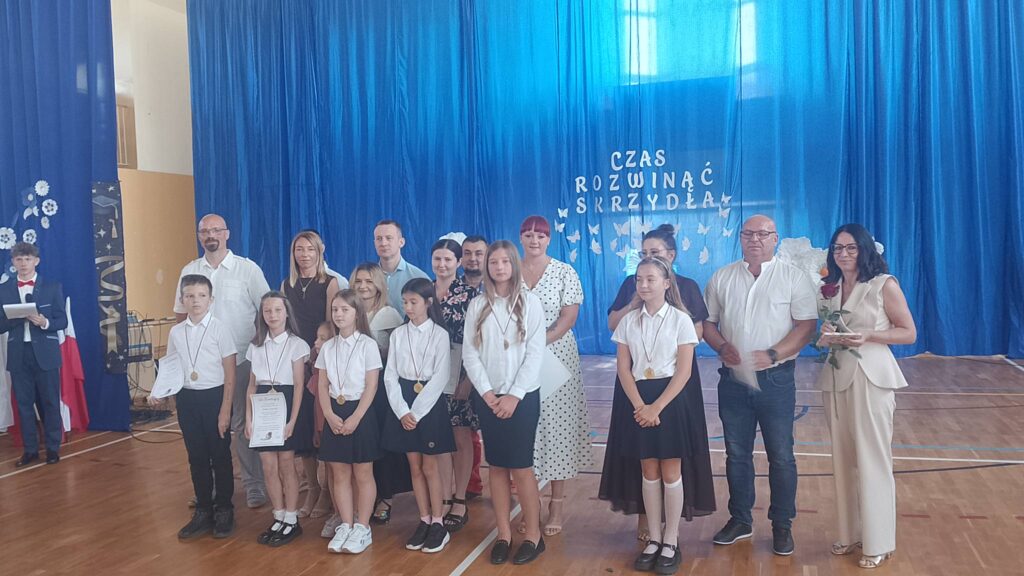 Na zdjęciu przedstawiono grupę uczniów wraz z rodzicami i opiekunami podczas uroczystości zakończenia roku szkolnego. Fotografia została wykonana w szkolnej sali gimnastycznej, na tle niebieskiej kurtyny z dekoracją i napisem „Czas rozwinąć skrzydła”, który symbolizuje rozwój oraz rozpoczęcie kolejnego etapu edukacji.

Na pierwszym planie stoją uczniowie ubrani w odświętne stroje – białe koszule i bluzki oraz ciemne spódnice lub spodnie. Na szyjach mają zawieszone złote medale, będące wyróżnieniem za osiągnięcia w nauce, sporcie lub działalności szkolnej. Dwoje uczniów trzyma w dłoniach pamiątkowe dyplomy potwierdzające otrzymane nagrody.
