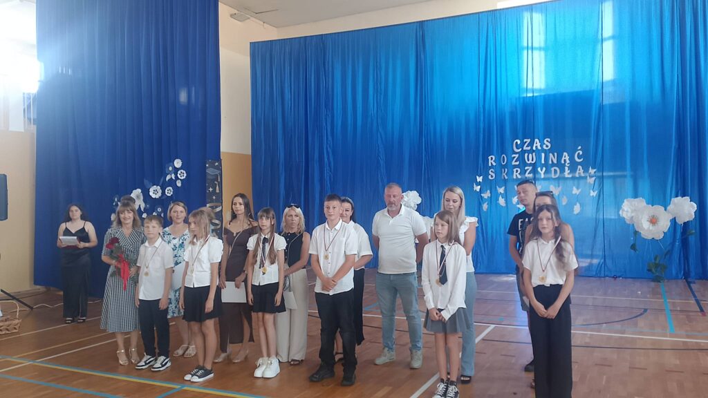 Na zdjęciu przedstawiono uczniów wraz z rodzicami i nauczycielami podczas uroczystości zakończenia roku szkolnego, pozujących do pamiątkowej fotografii po wręczeniu nagród i wyróżnień. Fotografia została wykonana w szkolnej sali gimnastycznej, której tło stanowi niebieska kurtyna z dekoracją i napisem „Czas rozwinąć skrzydła”, symbolizującym rozwój uczniów i rozpoczęcie nowego etapu edukacji.

Na pierwszym planie stoją wyróżnieni uczniowie ubrani w odświętne stroje – białe koszule i bluzki oraz ciemne spodnie lub spódnice. Na szyjach mają zawieszone medale będące wyrazem uznania za osiągnięcia w nauce, sporcie lub działalności szkolnej. Niektórzy z nich trzymają w dłoniach dyplomy, które otrzymali podczas uroczystości.