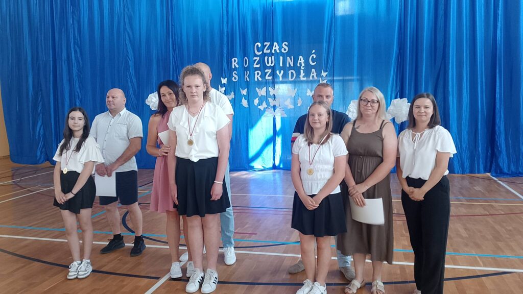 Na zdjęciu przedstawiono uczniów wraz z rodzicami i nauczycielami podczas uroczystości zakończenia roku szkolnego, pozujących do pamiątkowej fotografii po wręczeniu nagród i wyróżnień. Fotografia została wykonana w szkolnej sali gimnastycznej, której tło stanowi niebieska kurtyna z dekoracją i napisem „Czas rozwinąć skrzydła”, symbolizującym rozwój uczniów i rozpoczęcie nowego etapu edukacji.

Na pierwszym planie stoją wyróżnieni uczniowie ubrani w odświętne stroje – białe koszule i bluzki oraz ciemne spodnie lub spódnice. Na szyjach mają zawieszone medale będące wyrazem uznania za osiągnięcia w nauce, sporcie lub działalności szkolnej. Niektórzy z nich trzymają w dłoniach dyplomy, które otrzymali podczas uroczystości.