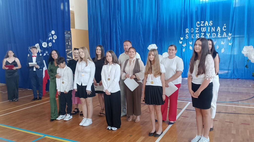 Na zdjęciu przedstawiono uczniów wraz z rodzicami i nauczycielami podczas uroczystości zakończenia roku szkolnego, pozujących do pamiątkowej fotografii po wręczeniu nagród i wyróżnień. Fotografia została wykonana w szkolnej sali gimnastycznej, której tło stanowi niebieska kurtyna z dekoracją i napisem „Czas rozwinąć skrzydła”, symbolizującym rozwój uczniów i rozpoczęcie nowego etapu edukacji.

Na pierwszym planie stoją wyróżnieni uczniowie ubrani w odświętne stroje – białe koszule i bluzki oraz ciemne spodnie lub spódnice. Na szyjach mają zawieszone medale będące wyrazem uznania za osiągnięcia w nauce, sporcie lub działalności szkolnej. Niektórzy z nich trzymają w dłoniach dyplomy, które otrzymali podczas uroczystości.