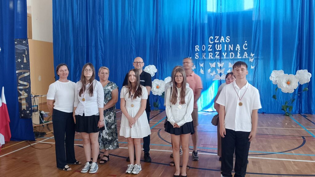 Na zdjęciu przedstawiono uczniów wraz z rodzicami i nauczycielami podczas uroczystości zakończenia roku szkolnego, pozujących do pamiątkowej fotografii po wręczeniu nagród i wyróżnień. Fotografia została wykonana w szkolnej sali gimnastycznej, której tło stanowi niebieska kurtyna z dekoracją i napisem „Czas rozwinąć skrzydła”, symbolizującym rozwój uczniów i rozpoczęcie nowego etapu edukacji.

Na pierwszym planie stoją wyróżnieni uczniowie ubrani w odświętne stroje – białe koszule i bluzki oraz ciemne spodnie lub spódnice. Na szyjach mają zawieszone medale będące wyrazem uznania za osiągnięcia w nauce, sporcie lub działalności szkolnej. Niektórzy z nich trzymają w dłoniach dyplomy, które otrzymali podczas uroczystości.