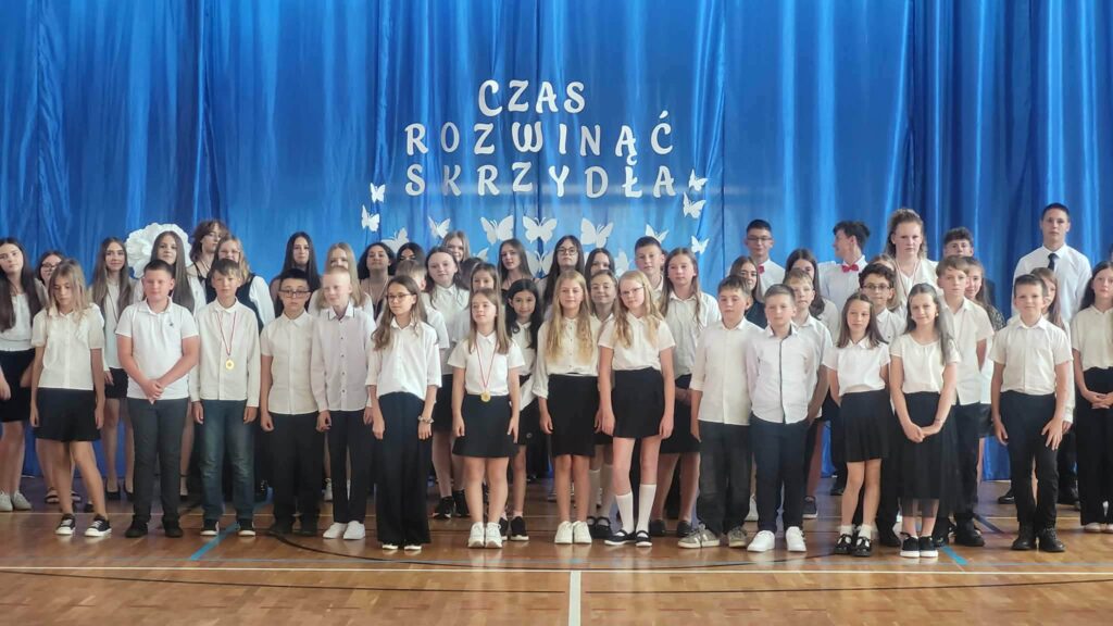 Na zdjęciu przedstawiono grupę uczniów zgromadzonych podczas uroczystości zakończenia roku szkolnego w szkolnej sali gimnastycznej. Wszyscy ustawieni są do wspólnego występu lub pamiątkowego zdjęcia na tle niebieskiej kurtyny z napisem „Czas rozwinąć skrzydła”, ozdobionej białymi motylami symbolizującymi rozwój i rozpoczęcie nowego etapu edukacji.

Na pierwszym planie stoją młodsi uczniowie ubrani w odświętne stroje – białe koszule lub bluzki oraz ciemne spodnie i spódnice. Kilkoro z nich ma na szyi medale, będące wyróżnieniem za osiągnięcia lub udział w konkursach. W drugim i trzecim rzędzie znajdują się starsi uczniowie, również ubrani elegancko, którzy wspólnie tworzą liczny zespół uczestniczący w akademii.