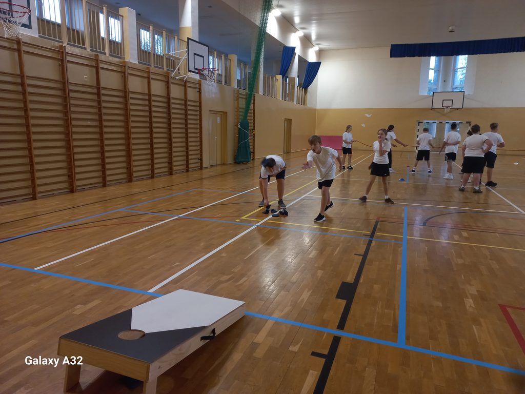 Na zdjęciu są uczniowie w białych koszulkach i ciemnych spodenkach, którzy grają w Cornhole. Znajdują się na sali gimnastycznej. Dwóch z nich rzuca woreczkami do celu na podłodze.
