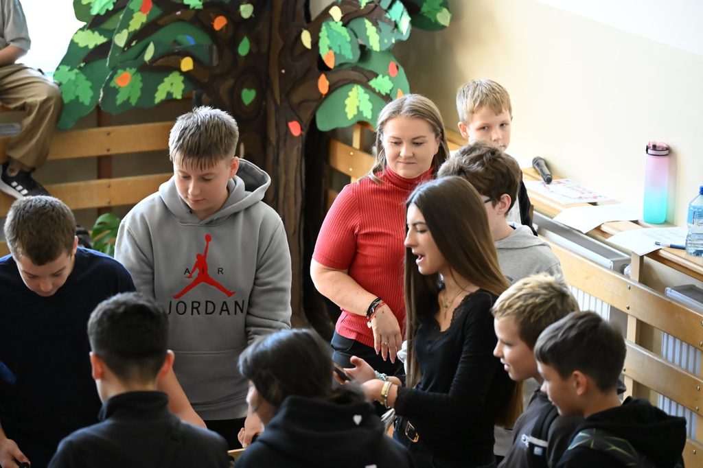Na zdjęciu widzimy grupę uczniów oraz osoby dorosłe uczestniczące we wspólnym wydarzeniu integracyjnym. Uczestnicy zgromadzili się wokół miejsca, w którym odbywa się wspólna aktywność. Rozmawiają, obserwują jej przebieg i angażują się w spotkanie.
Fotografia przedstawia przyjazną i pełną zaangażowania atmosferę. Obecność zarówno młodzieży, jak i dorosłych podkreśla wspólnotowy charakter wydarzenia. Takie spotkania są okazją do integracji, wzmacniania relacji, rozwijania umiejętności komunikacyjnych oraz wspólnego spędzania czasu. Kolorowa dekoracja w kształcie drzewa widoczna w tle dodatkowo nadaje przestrzeni ciepły i przyjazny charakter.