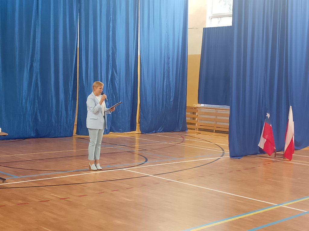Na zdjęciu widzimy uroczystość odbywającą się w sali gimnastycznej szkoły. Na pierwszym planie stoi kobieta ubrana w jasny garnitur. Trzyma w ręku mikrofon oraz przygotowany tekst i przemawia do zgromadzonych uczestników spotkania.
W tle znajdują się duże niebieskie kotary, które tworzą uroczystą oprawę wydarzenia. Po prawej stronie ustawiono obok siebie flagi Chile i Polski, symbolizujące spotkanie przedstawicieli obu krajów. Fotografia przedstawia najprawdopodobniej moment oficjalnego powitania gości z Chile.