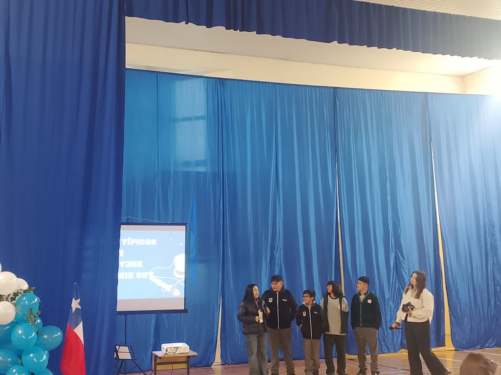 Na zdjęciu widzimy uczniów z Chile podczas prezentacji przygotowanej dla społeczności szkolnej. Grupa stoi przed niebieską kotarą, a jedna z osób dorosłych towarzyszy uczniom podczas wystąpienia.
Po lewej stronie znajduje się ekran, na którym wyświetlana jest przygotowana prezentacja. Obok widoczna jest flaga Chile oraz dekoracja z biało-niebieskich balonów, podkreślająca uroczysty charakter spotkania.
Podczas wystąpienia uczniowie z Chile przedstawiali siebie, opowiadali o swoich zainteresowaniach, codziennym życiu oraz kulturze swojego kraju.