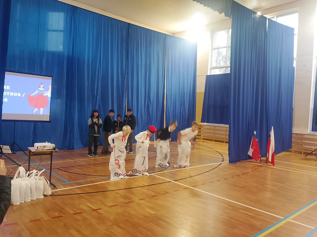 Na zdjęciu widzimy uczniów podczas wspólnej zabawy przygotowanej w ramach spotkania z gośćmi z Chile. Na pierwszym planie cztery osoby uczestniczą w konkurencji polegającej na poruszaniu się w dużych materiałowych workach. W tle obserwują ich uczniowie z Chile.
Po lewej stronie znajduje się ekran z prezentacją, natomiast po prawej ustawiono flagi Chile i Polski, które podkreślają międzynarodowy charakter wydarzenia.
Fotografia pokazuje radosny i integracyjny moment spotkania. Wspólne zabawy były doskonałą okazją do poznania tradycji, przełamania barier językowych oraz budowania przyjaznych relacji między uczniami z Polski i Chile.