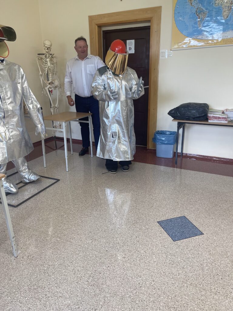 Zdjęcie przedstawia pracownika Huty oraz uczennicę klasy 8 ubraną w odzież ochronną (z zamkniętą przyłbicą na kasku ochronnym), która jest obowiązkowa w czasie pracy przy piecach hutniczych
