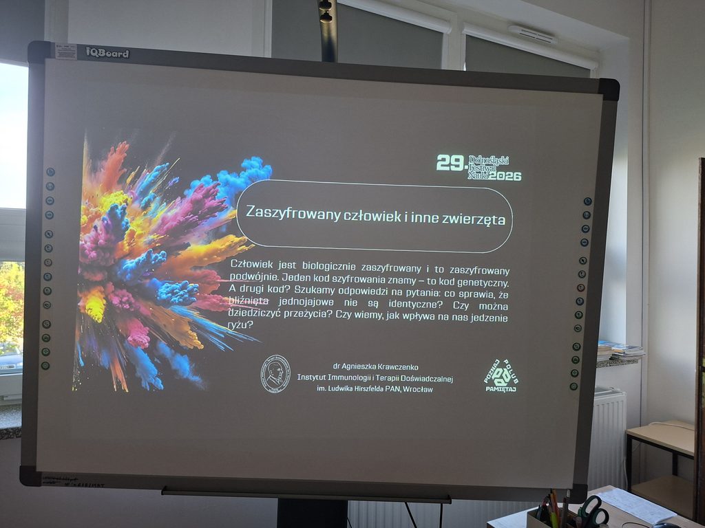 Przedstawia ekran multimedilany na którym wyświetlony jest tytuł spotkania "Zaszyfrowany człowiek i inne zwierzęta" w ramach Dolnośląskiego Festiwalu Nauki. Na ekranie znajdują się kolorowe elementy eksperymentów chemicznych, które symbolizują wielokierunkowość nauki. Tekst jest napisany białą czcionką na czarnym tle. 
