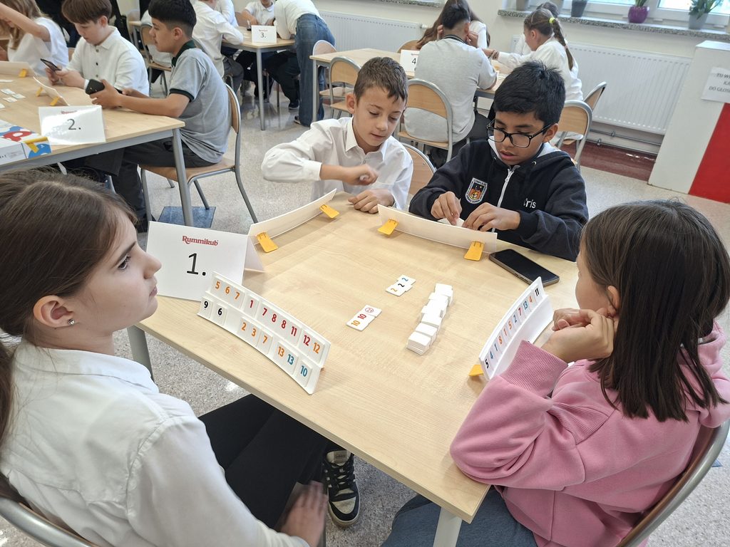 djęcie przedstawia uczniów grających w Rummikub. Dzieci siedzą na krzesłach a na stolikach są klocki z gry Rummikub, które zawodnicy układają na podstawkach, a następnie poprawnie ułożone sekwencje wykładają na stół.