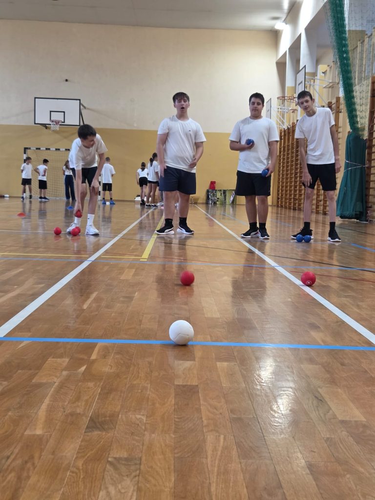 Na zdjęciu widzimy uczniów aktywnie uczestniczących w sportowych zmaganiach na sali gimnastycznej. Chłopcy biorą udział w konkurencji zręcznościowej z wykorzystaniem kolorowych piłeczek. Zadanie wymaga od uczestników celności, skupienia, precyzji i koordynacji ruchowej.
W tle pozostali uczniowie również uczestniczą w sportowych aktywnościach.