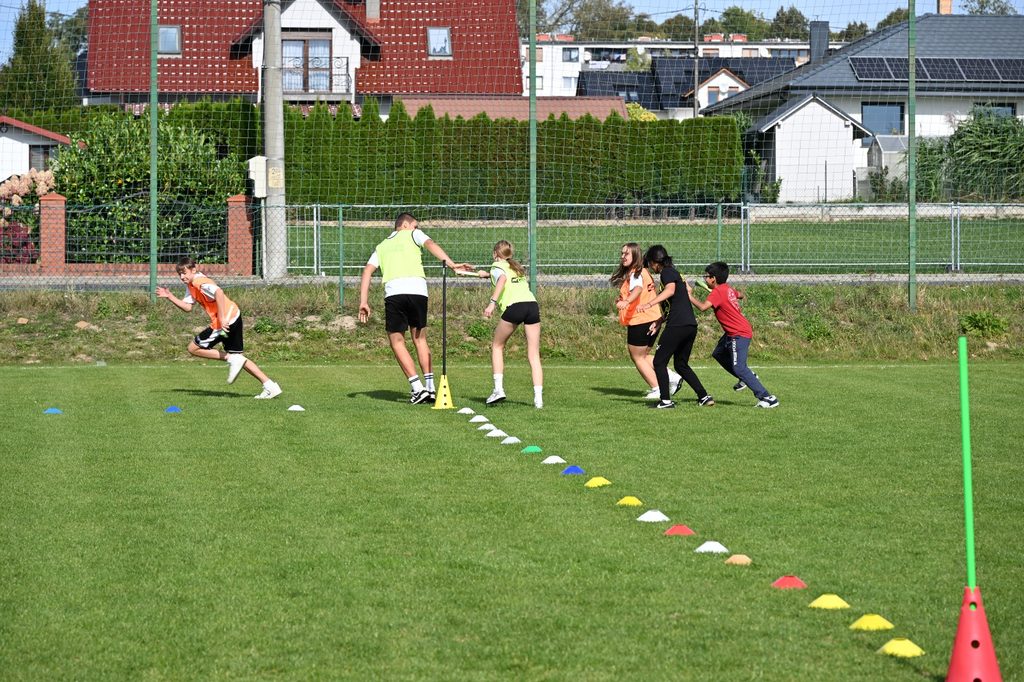 Na zdjęciu widzimy uczniów podczas sportowej rywalizacji w ramach „Porowej sztafety”. Zawodnicy z dużym zaangażowaniem pokonują wyznaczoną trasę na szkolnym boisku, oznaczoną kolorowymi pachołkami. Uczestnicy czekają na swoją kolej, a kolejni zawodnicy ruszają do biegu.
Fotografia pokazuje ruch, emocje i ducha zdrowej rywalizacji.