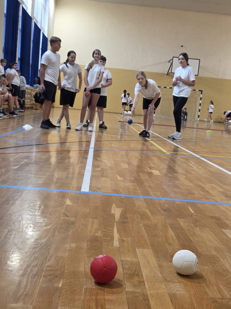 Na zdjęciu widzimy uczniów uczestniczących w emocjonującej konkurencji sportowej na sali gimnastycznej. Tym razem zadaniem uczestników jest precyzyjne toczenie kolorowych piłeczek w kierunku wyznaczonego celu.
Jedna z uczennic właśnie wykonuje swój rzut, a pozostali zawodnicy z zainteresowaniem obserwują jej próbę i czekają na swoją kolej.
Ta konkurencja to doskonały sposób na rozwijanie celności, koncentracji, koordynacji ruchowej oraz umiejętności zdrowej rywalizacji. Nie zabrakło sportowych emocji, wzajemnego wsparcia i dobrej zabawy!