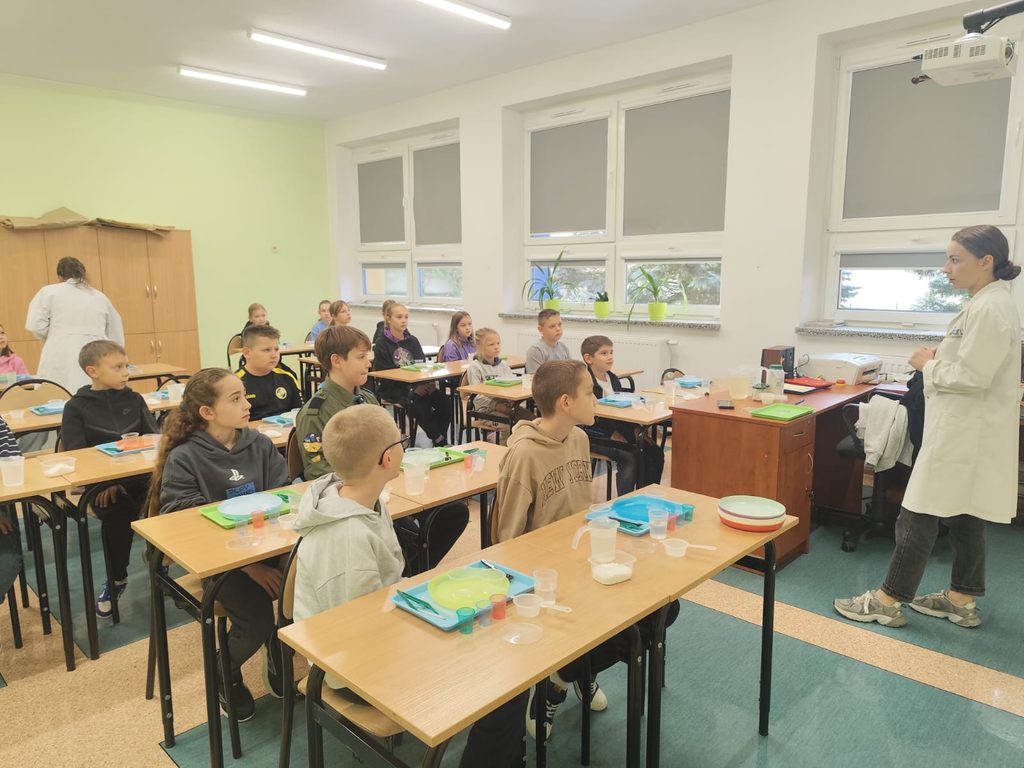 Na zdjęciu widzimy salę lekcyjną, w której odbywają się zajęcia edukacyjne lub warsztaty praktyczne dla dzieci.
Uczniowie siedzą w szkolnych ławkach i z zainteresowaniem słuchają prowadzącej. Kobieta ubrana w biały fartuch stoi przed klasą i prawdopodobnie wyjaśnia zasady wykonywania doświadczenia lub zadania.
Na ławkach znajdują się kolorowe tacki, plastikowe kubeczki, łyżeczki, talerzyki oraz różne materiały potrzebne do przeprowadzenia eksperymentów.