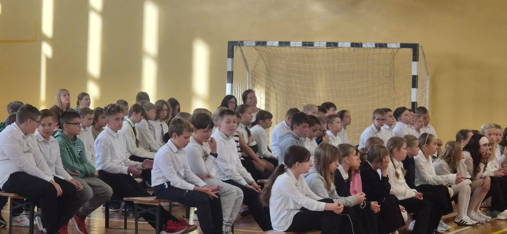 Na zdjęciu przedstawiono uczniów uczestniczących w uroczystości szkolnej odbywającej się w sali gimnastycznej. Dzieci i młodzież, ubrane w eleganckie, odświętne stroje, zajmują miejsca na ławkach i z uwagą obserwują przebieg wydarzenia.
Wśród zgromadzonych widoczni są uczniowie w różnym wieku, a także nauczyciele i pozostali uczestnicy uroczystości. Wspólne spotkanie stanowi okazję do integracji społeczności szkolnej, kształtowania właściwych postaw oraz rozwijania umiejętności uczestniczenia w wydarzeniach o charakterze kulturalnym i wychowawczym.
