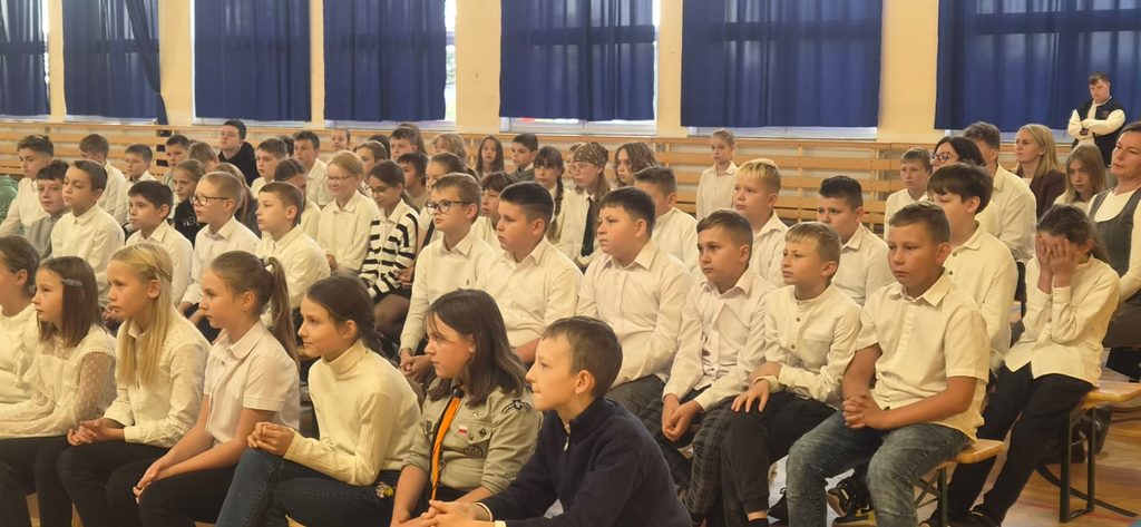 Na zdjęciu przedstawiono uczniów uczestniczących w uroczystości szkolnej odbywającej się w sali gimnastycznej. Dzieci i młodzież, ubrane w eleganckie, odświętne stroje, zajmują miejsca na ławkach i z uwagą obserwują przebieg wydarzenia.
Wśród zgromadzonych widoczni są uczniowie w różnym wieku, a także nauczyciele i pozostali uczestnicy uroczystości. Wspólne spotkanie stanowi okazję do integracji społeczności szkolnej, kształtowania właściwych postaw oraz rozwijania umiejętności uczestniczenia w wydarzeniach o charakterze kulturalnym i wychowawczym.