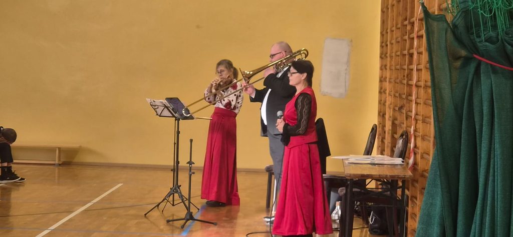 Na zdjęciu przedstawiono występ artystyczny zespołu muzycznego w sali gimnastycznej. Dwoje muzyków gra na instrumentach dętych blaszanych, a towarzysząca im wokalistka trzyma mikrofon. Wykonawcy prezentują się w eleganckich strojach, a występ odbywa się przy akompaniamencie instrumentów na żywo.