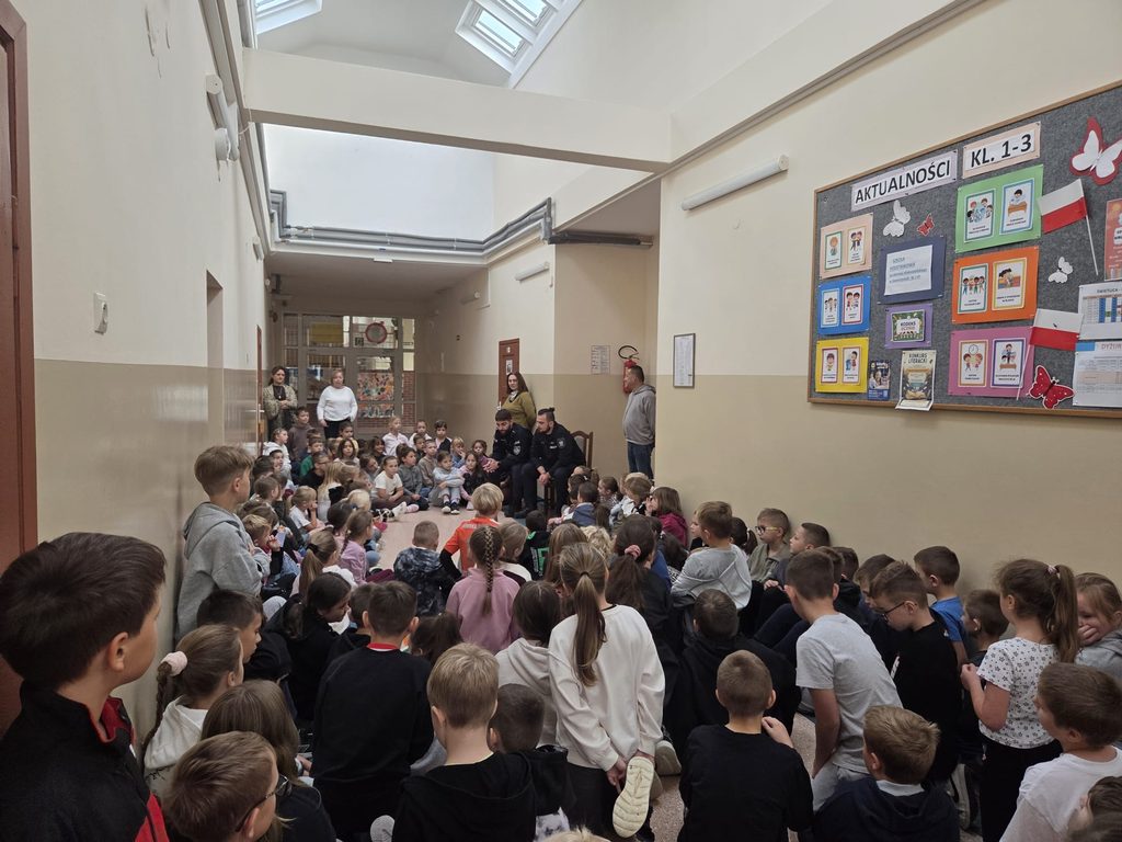 Na zdjęciu widać grupę dzieci z klas 1–3, które uczestniczą w spotkaniu z funkcjonariuszami policji. Dzieci siedzą na podłodze i z zainteresowaniem słuchają prelekcji dotyczącej bezpieczeństwa. Policjanci siedzą na krzesłach i przekazują uczniom ważne informacje. Korytarz jest jasny i przestronny, na ścianach znajdują się tablice informacyjne,