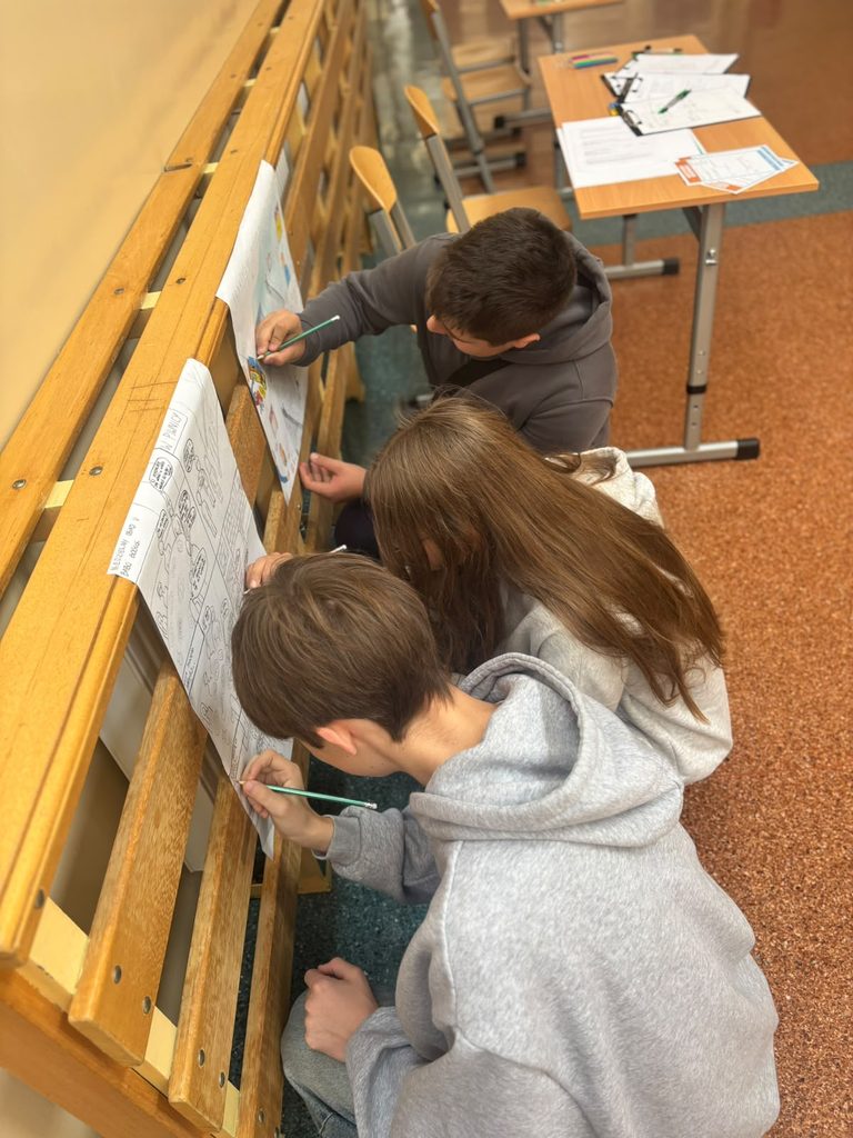 Na zdjęciu przedstawiono uczniów zaangażowanych w przygotowywanie plakatów edukacyjnych dotyczących bezpieczeństwa. Dzieci wspólnie pracują nad ilustracjami, wykorzystując kredki i własne pomysły do przedstawienia zasad bezpiecznego zachowania.
Działanie wpisuje się w tematykę Krajowego Treningu Rodzinnego – Akademii Ochrony Ludności i Obrony Cywilnej (OLiOC). Poprzez aktywność plastyczną uczniowie utrwalają wiedzę na temat bezpieczeństwa, rozwijają kreatywność, umiejętność współpracy oraz poczucie odpowiedzialności za siebie i innych. 