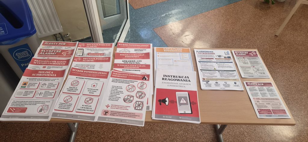 Na zdjęciu przedstawiono materiały edukacyjne przygotowane w ramach Krajowego Treningu Rodzinnego – Akademii Ochrony Ludności i Obrony Cywilnej (OLiOC).
Na stole wyeksponowano plakaty, ulotki i instrukcje dotyczące bezpieczeństwa oraz właściwego postępowania w sytuacjach zagrożenia. Materiały obejmują m.in. zasady reagowania na sygnały alarmowe i Alerty RCB, wybór bezpiecznego miejsca schronienia, przygotowanie rodzinnego planu bezpieczeństwa oraz postępowanie w przypadku zagrożenia atakiem z powietrza.