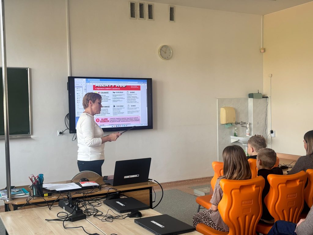 Na zdjęciu przedstawiono zajęcia edukacyjne przeprowadzone w ramach Krajowego Treningu Rodzinnego – Akademii Ochrony Ludności i Obrony Cywilnej (OLiOC).
Podczas spotkania uczniowie uczestniczyli w prelekcji poświęconej zasadom bezpieczeństwa oraz właściwego reagowania w sytuacjach zagrożenia. Prowadząca, wykorzystując prezentację multimedialną, omawiała zagadnienia związane z Alertami RCB, sygnałami alarmowymi oraz postępowaniem w przypadku zagrożenia atakiem z powietrza.