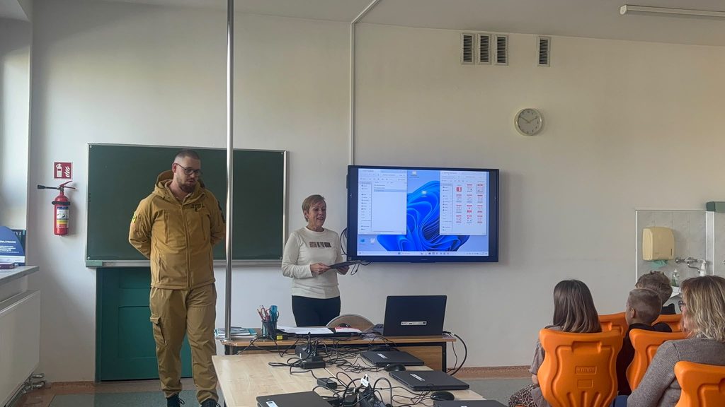 Na zdjęciu przedstawiono spotkanie edukacyjne z udziałem przedstawiciela służb mundurowych, zorganizowane w ramach Krajowego Treningu Rodzinnego – Akademii Ochrony Ludności i Obrony Cywilnej (OLiOC).
W szkolnej sali prowadząca spotkanie oraz zaproszony gość w umundurowaniu uczestniczą w zajęciach poświęconych bezpieczeństwu i przygotowaniu do sytuacji kryzysowych. W spotkaniu biorą udział dzieci i dorośli, którzy mają okazję poszerzyć swoją wiedzę na temat ochrony życia i zdrowia, właściwego reagowania na zagrożenia oraz znaczenia odpowiedzialnego zachowania w sytuacjach niebezpiecznych.